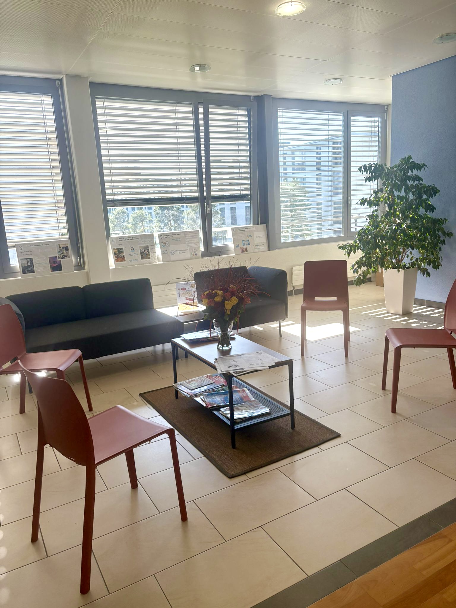 Intérieur d'un espace d'attente moderne avec des canapés, des chaises rouges et une table basse, baigné de lumière naturelle grâce aux grandes fenêtres.